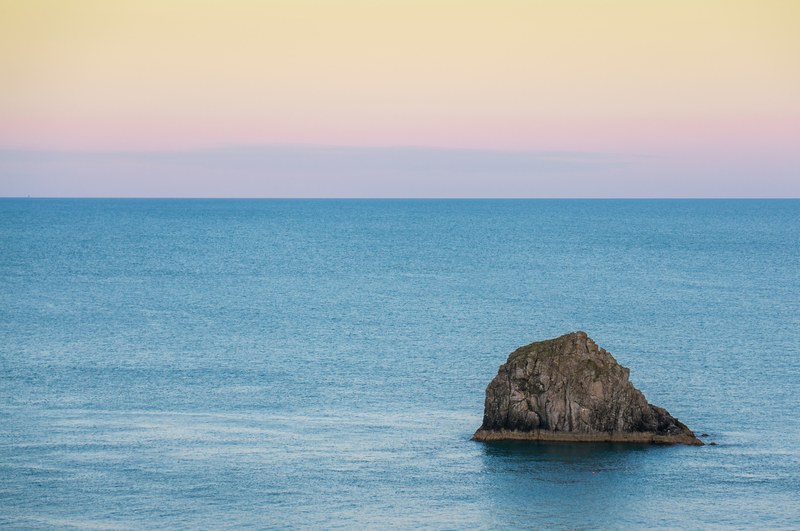 a lone rock formation protruding from the azure sea under a pale orange sky, lone rock in the sea 4k