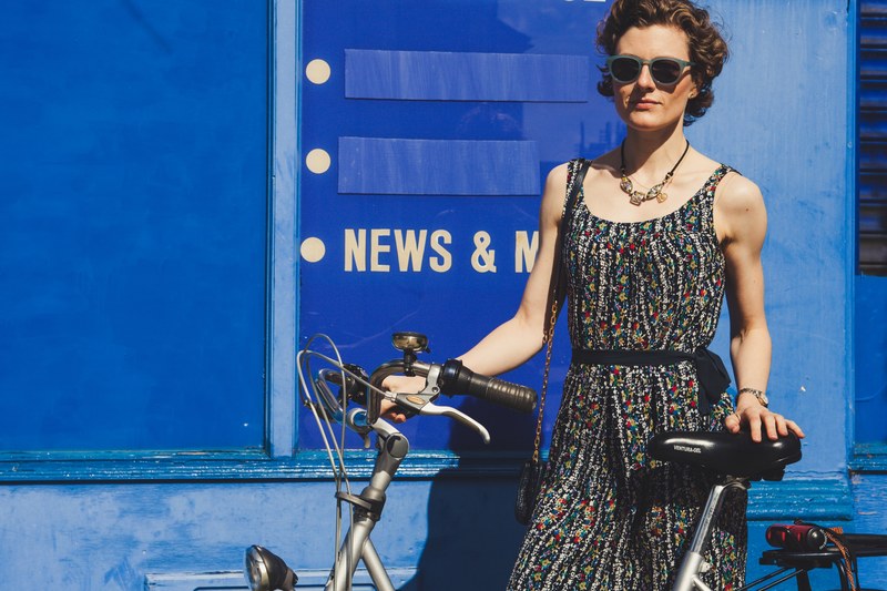 a woman wearing a floral print dress on a sunny day standing against a blue wall holding her bicycle in london, london bike 4k