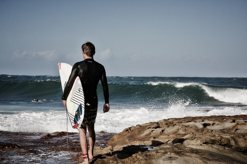 a guy wearing a wetsuit holding a surfboard looking out into the waves at dee why, over the rocks to the surf 4k