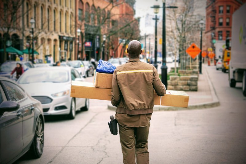 a bald man in uniform is carrying parcels next to a busy street in st lawrence market, _doing his job 4k