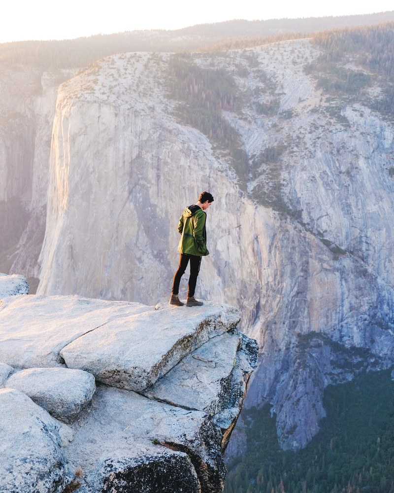 a man wearing black pants and a green jacket leaning over the edge of a cliff in yosemite valley, yosemite valley cliff 4k