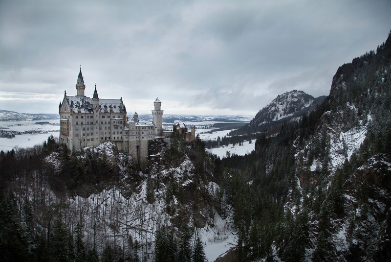 a massive white german castle sits among snowy forest trees and next to the foot of a mountain, neuschwansteinstrae castle 4k