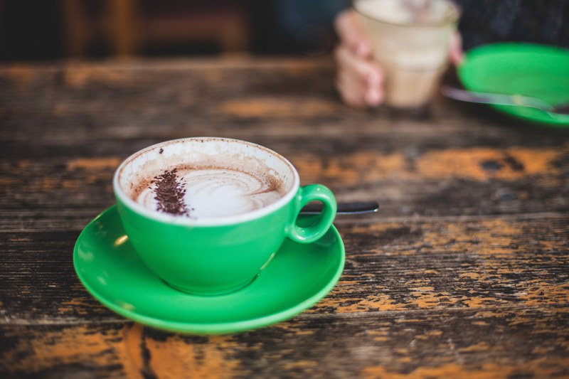 a latte in a green cup on a table with a person drinking their coffee on the other side, frothy coffee in a green cup 4k