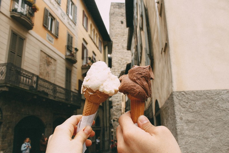 man and woman hold up chocolate and vanilla ice cream cones in old side street, outside the ice cream shop 4k