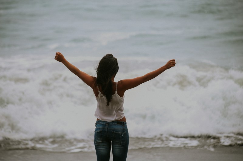 a woman wearing jeans and a white shirt staring out at the ocean with her arms up at bodega bay, bodega bay ocean 4k