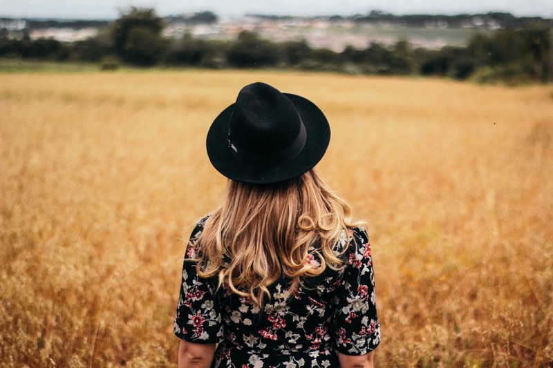 behind a woman standing alone in a wheat field waring a hat and dress, standing in a field 4k