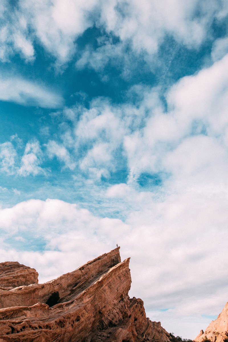 a distant silhouette on a hiker on top of a slanted orange rock ledge under a cloudy azure sky, hiker on a steep orange ledge 4k