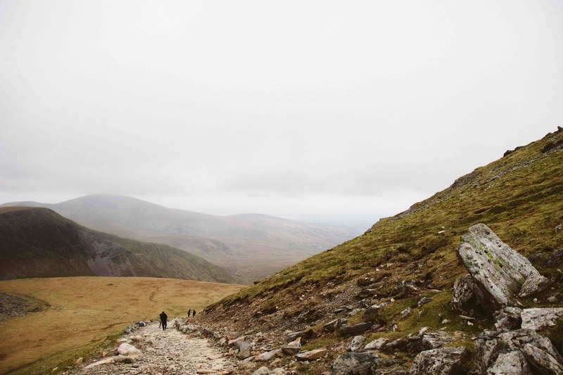 hikers climbing up a rocky path at the foot of a green hill on a cloudy day, hiking up the hills 4k