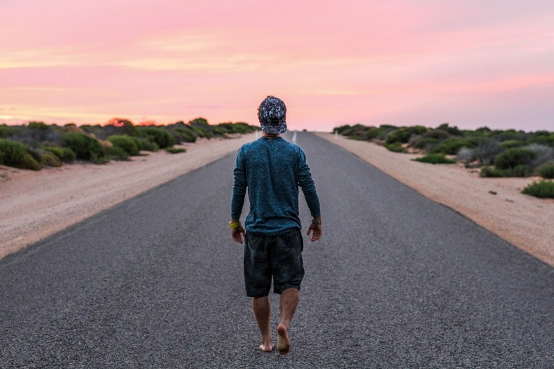 a barefoot man in a baseball hat walking on an empty asphalt road during sunset, lone man on an asphalt road 4k