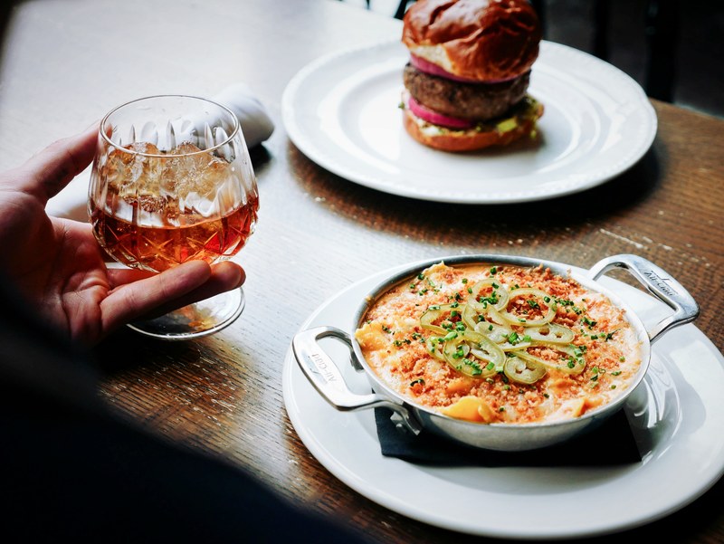 hand holding a cocktail next to a cheese and jalapeno dip and a burger, happy hour food and drinks 4k