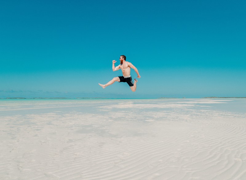 a shirtless muscular man in shorts in the middle of a high jump above a sandy beach, sandbar jump 4k