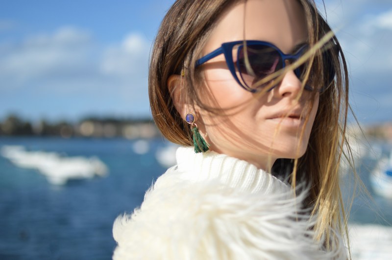 a faint smile on the face of a young woman in sunglasses over a body of water, faux fur fashion 4k
