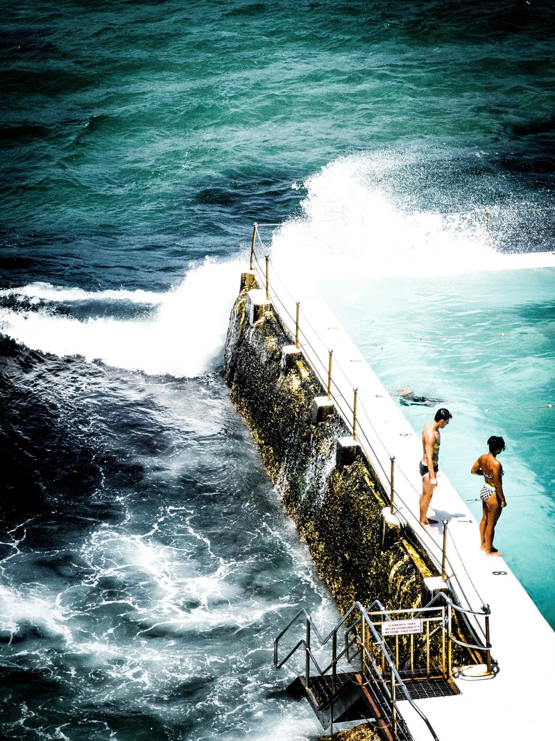a couple standing on the step of an outdoor pool to swim in bondi icebergs pool, a couple standing by pool step 4k