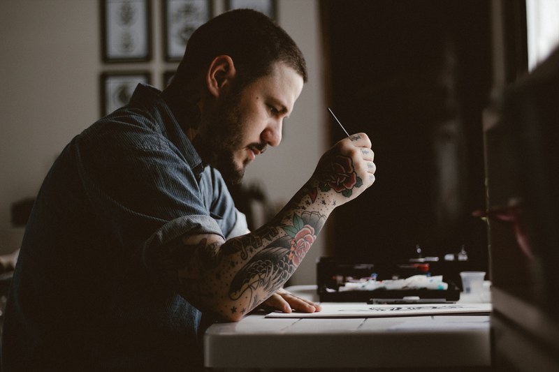 an artist with a tattoo on his arm working at a desk, tattooed artist at work 4k