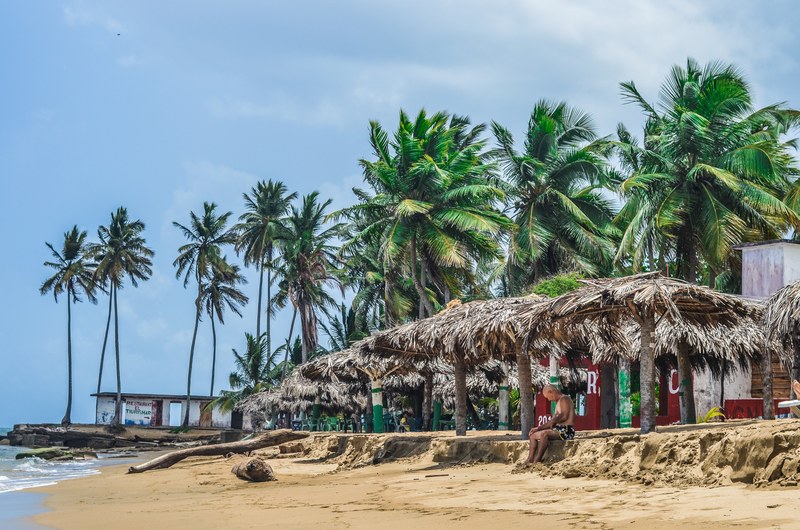 a lone man sitting on the beach under a thatched roof, sheltering from sun on a beach 4k