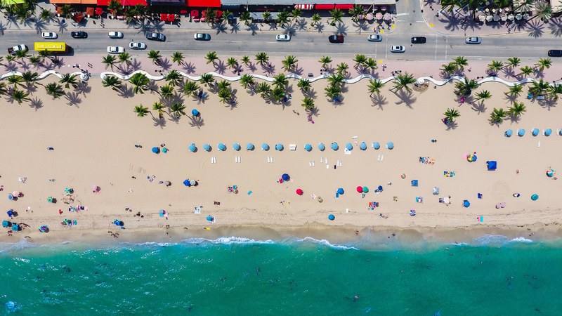 a drone shot of people vacationing on a beach lined with palm trees and blue umbrellas, floridas streets 4k