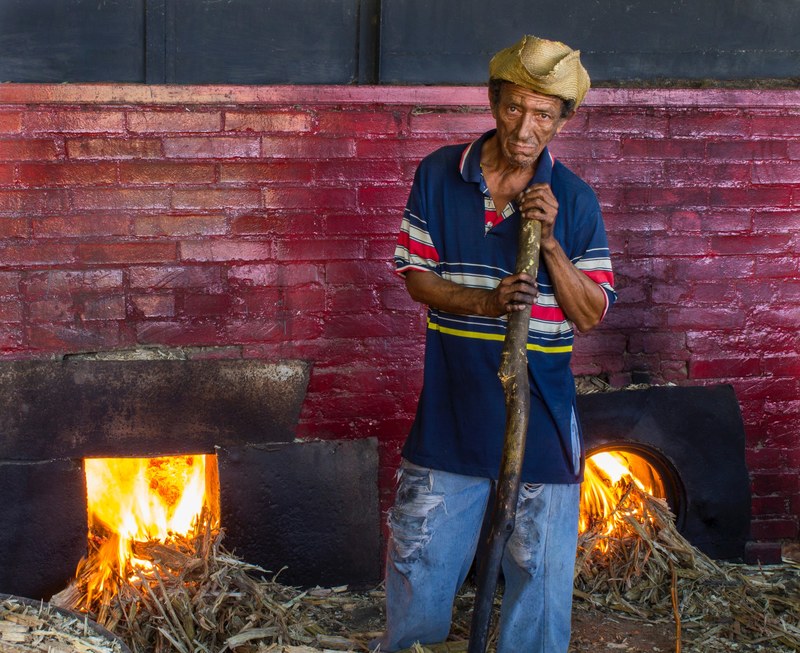 a serious worker in a striped shirt straw hat and distressed jeans leaning on a wooden stick blackened by soot burning wood in two red brick fireplaces, trapiche en curarigua 4k