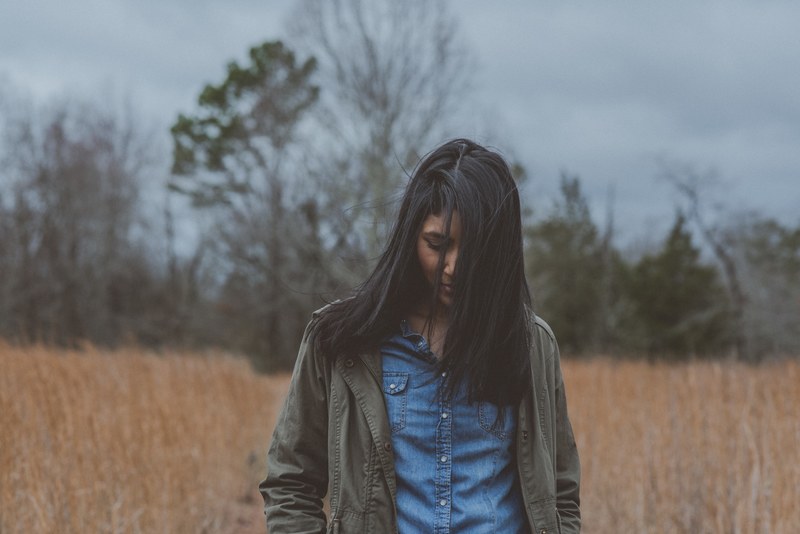 a woman with black hair and a jacket looks down in a field near trees, whats she thinking 4k