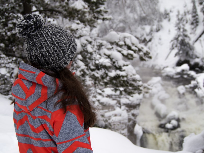 a woman wearing a gray and red hoodie and a beanie cap staring through a wintery forest, waterfall view 4k