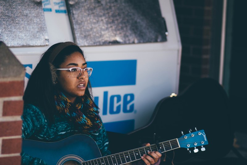 a woman in glasses and earmuffs playing a blue acoustic guitar near a building, cedarville part iii 4k