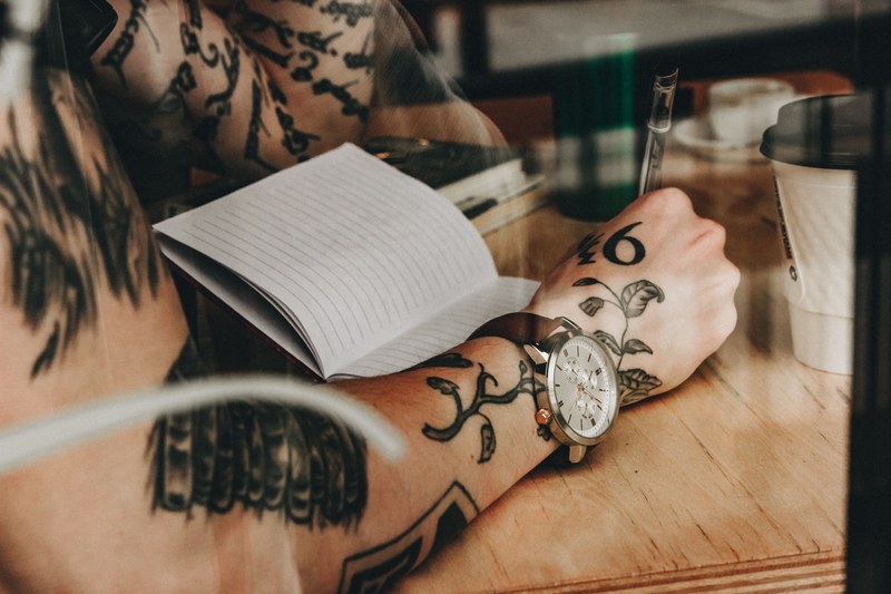 milwaukee man with tattoos wearing wristwatch writing in journal while sitting at cafe, captivated_ 4k