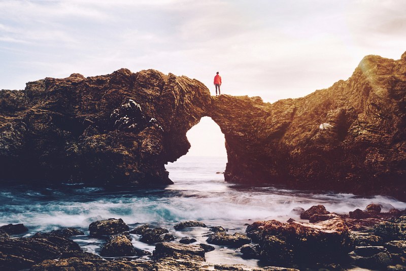 a breathtaking shot of a person standing on an arch created by a jagged cliff over water, looking off into the sea 4k
