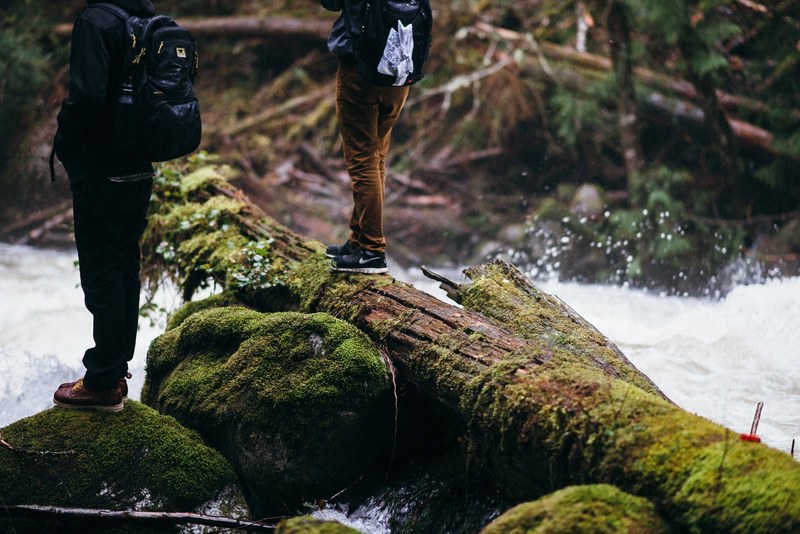 two people on mossy rocks and logs in a fast flowing river, two men over a frothy river 4k
