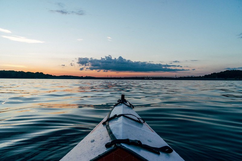 a perspective shot from a kayak on a rippling lake with the sun setting on the horizon in the distance, kayaking at sunset 4k