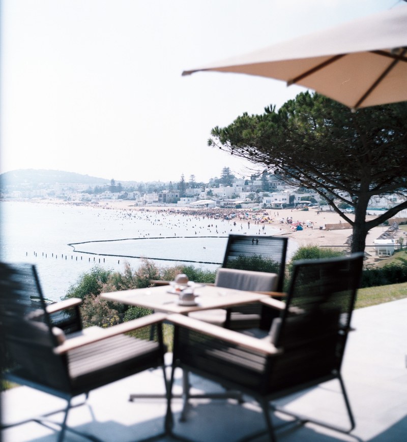 a patio with three chairs at a square table with a view on a crowded beach and a coastal town, au calme 4k
