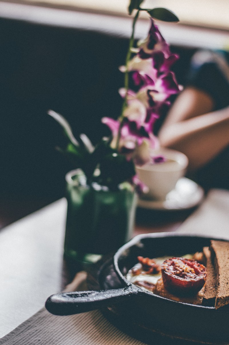 a blurry shot of a skillet with a fried tomato and bread next to a flower vase on a table, flowery breakfast 4k