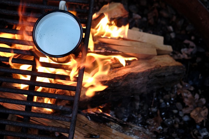 a tin cup of water on a grill above a campfire made of wooden logs in a fire pit, tea time 4k