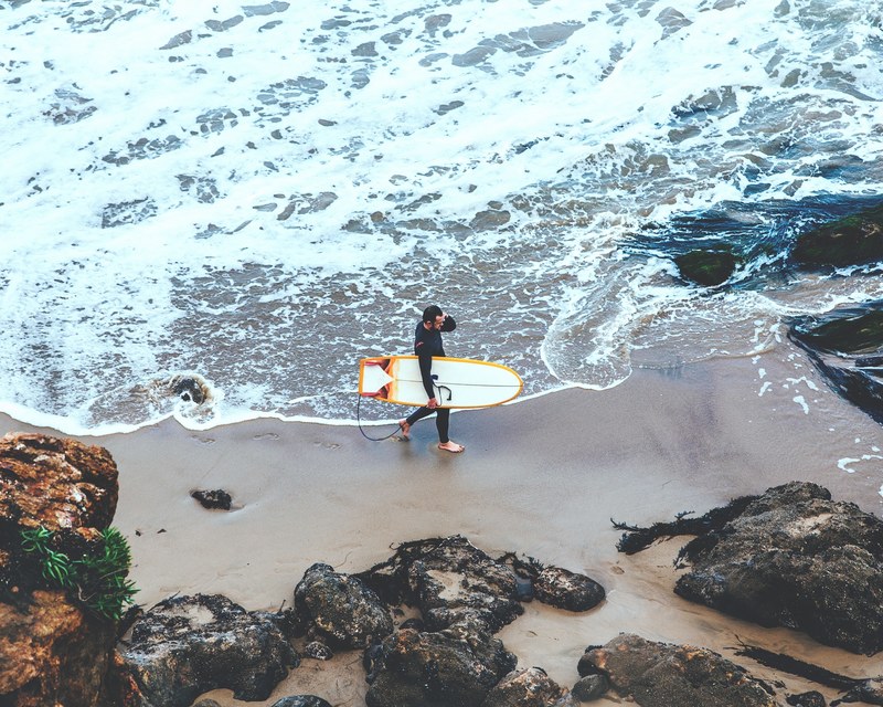 a shot of a surfer carrying a white board along a small bay taken from above as waves lap the shore at point dume, surfer below carrying his board 4k
