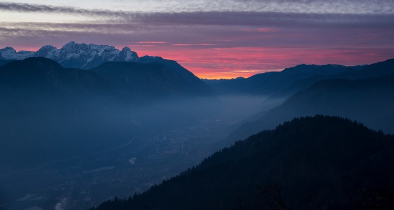 an aerial view in ajdna as a mist settles into the valley_ the sunrise or sunset behind the distant snowcap mountains turn the cloudy sky orange and pink, dark sunset 4k