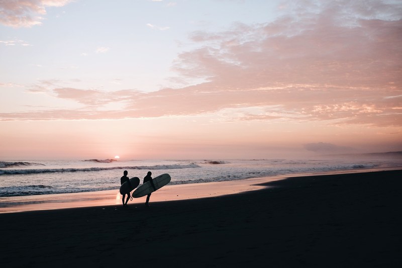 silhouettes of two people carrying their surfboards on an empty beach during sunset, surfer silhouettes at dusk 4k