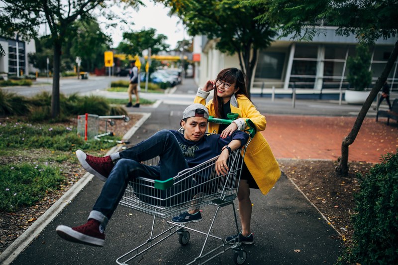 a woman leans onto a shopping cart while a man poses inside on a city sidewalk, posing with shopping cart 4k