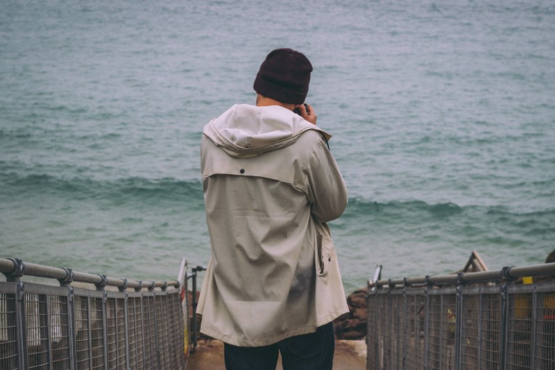 a man wearing a fall jacket on a fenced path walking toward the sea with a camera, plmont ocean photography 4k