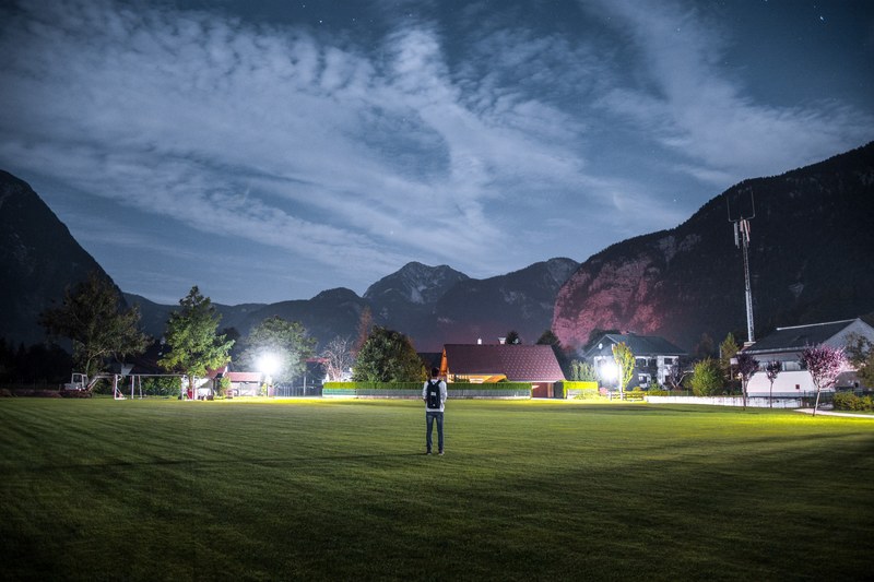 man with a backpack standing on the soccer field in obertraun at night, man standing on the field at night 4k