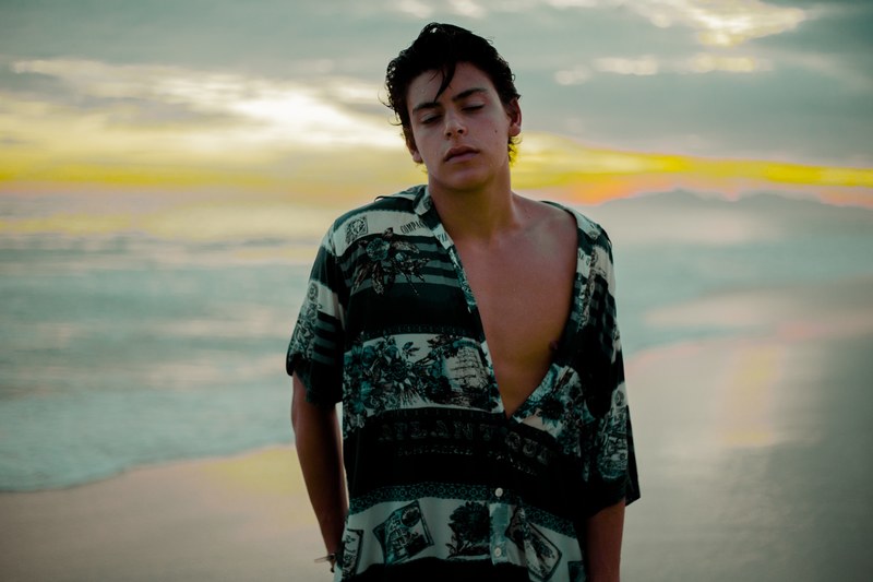 young man standing on praia da barra da tijuca beach with waves crashing and a sunset behind him, man at praia da barra da tijuca 4k