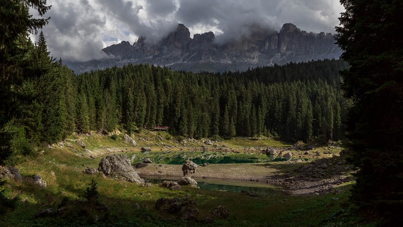 a small lake surrounded by rocks and coniferous trees with tall mountains on the horizon, lake near the mountains in carezza 4k