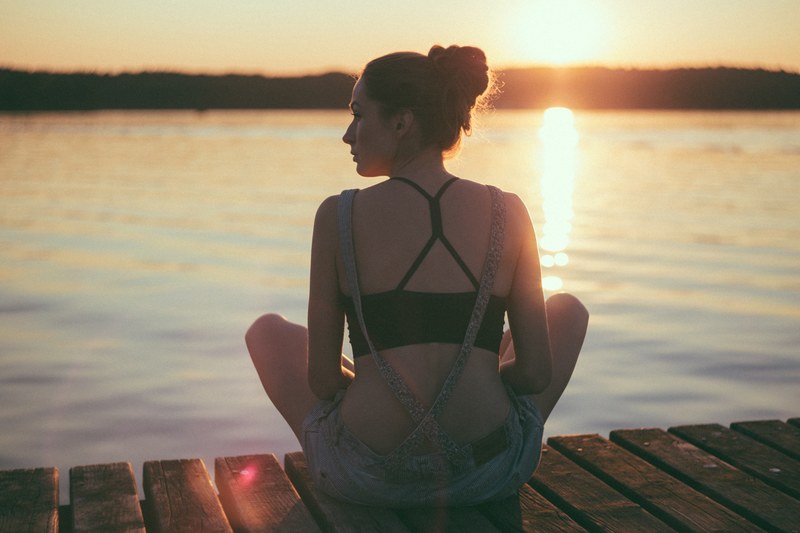 woman in dungarees sitting on dock looking to the left whil sun sets in the horizon and reflects over the water in olsztyn, woman on dock sunset 4k