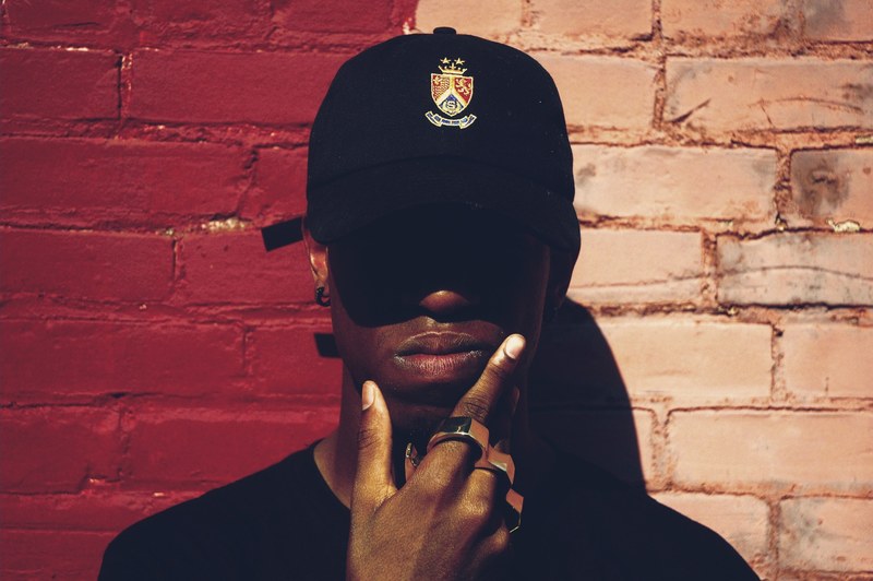 a man wearing a baseball cap brass knuckles and earrings holding his chin by a brick wall, man by a brick wall 4k