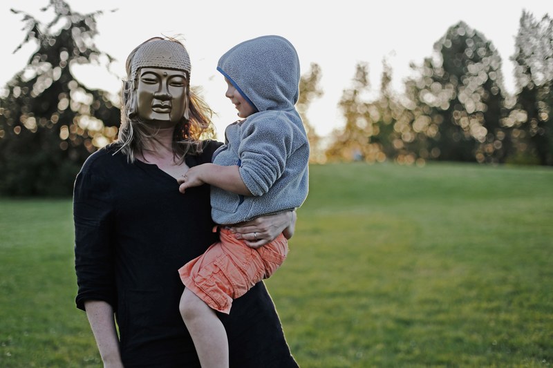 a woman wearing a buddha mask is carrying a child wearing a hooded jacket, _buddha in the field 4k
