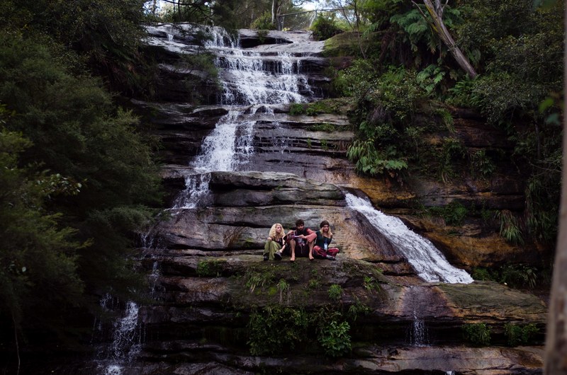 three young people sitting on a rocky ledge with a waterfall streaming down on their side, sitting on a waterfall ledge 4k