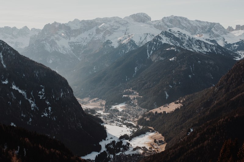 a mountain valley covered in dark trees and a town at the foot of the mountain covered in snow, mountain valley in canazei 4k