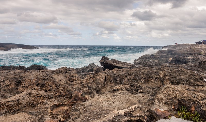 azure waves crash against the rocky shore as two silhouettes sit with fishing rods in the distance, fishing on the rocky shore 4k