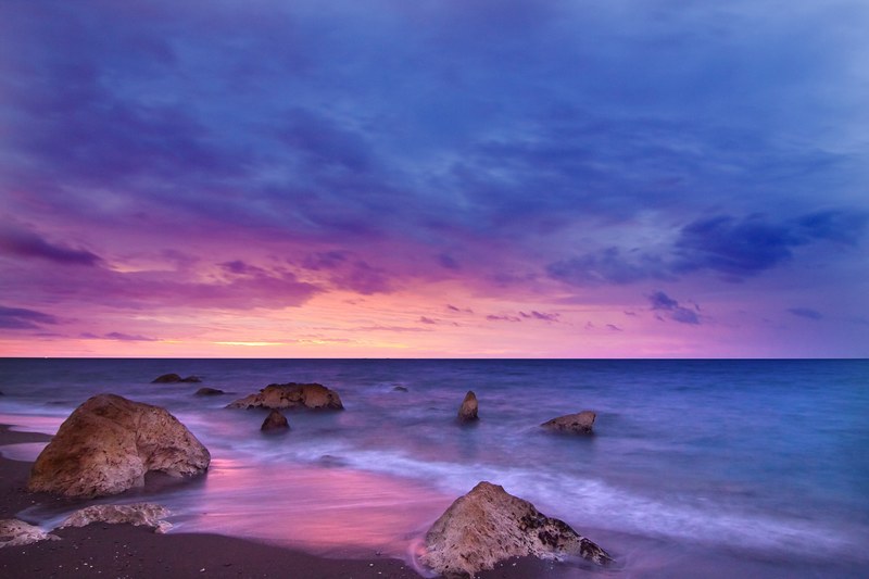 rocks sit in a calm sea at twilight as the sunset creates a pink purple and navy sky at playa del penn del cuervo, playa del penn del cuervo sunset_ 4k