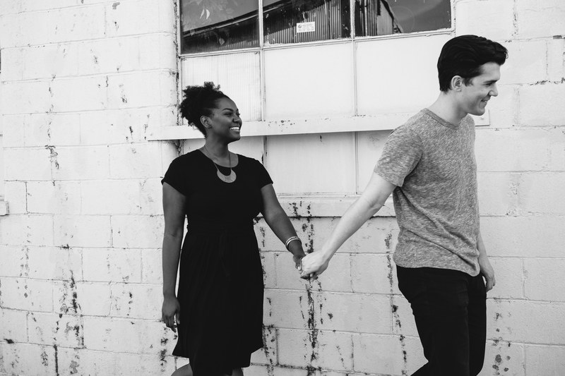 a couple in black and white smiles and holds hands aside a white brick wall, couple holding hands by a wall 4k
