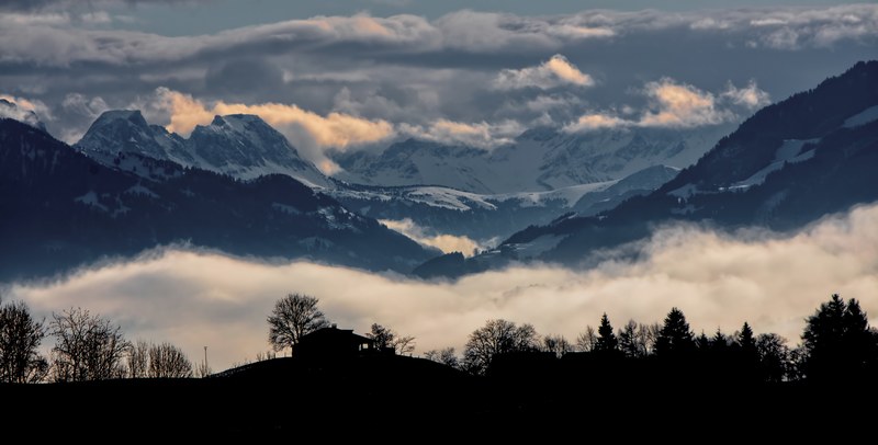 dark silhouettes of a house and trees against white clouds in a snowy mountain valley, stunning view 4k