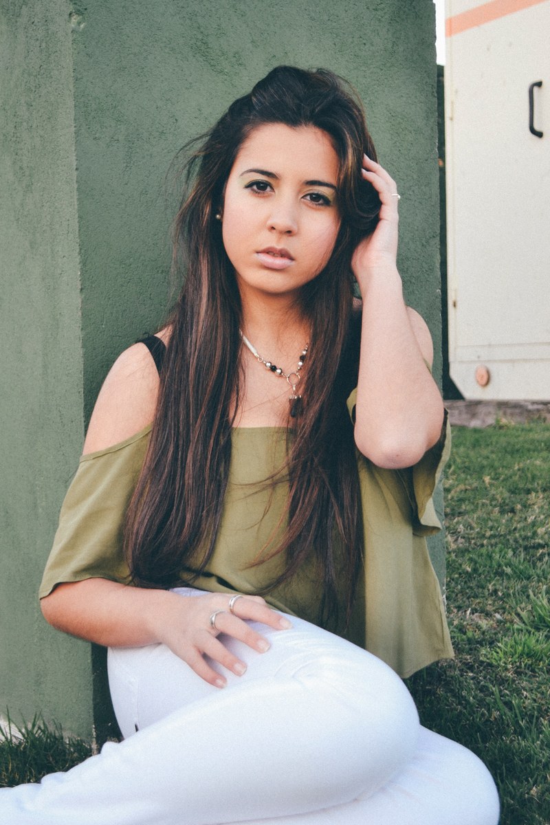 a woman with long dark brown hair sits in the grass and poses by putting on hand one her thigh and the other in her hair, woman sitting on grass 4k