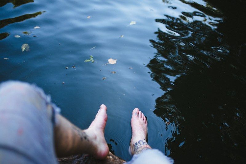 man with tattooed legs dangling his feet above the reflective lake waters of abbots pool, man relaxing feet in pool 4k